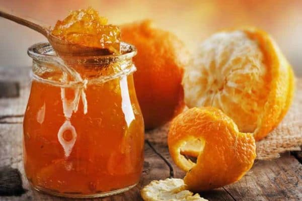 Orange jam in a glass scooped with a spoon and a half-peeled orange is placed next to it.