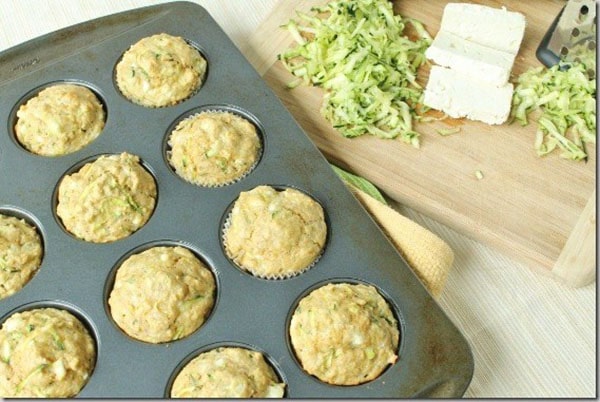 Zucchini muffins in a muffin tin and a wooden board with grated zucchini is placed next to it.
