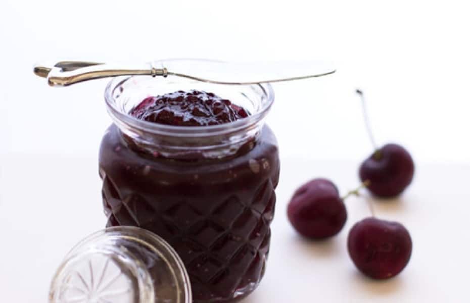 Marmeláda z višní ve sklenici, na které je položen nůž a vedle jsou pohozeny čerstvé višně.