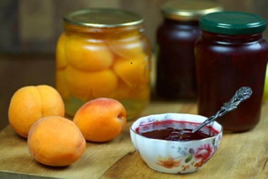 Pickled apricots in jars with fresh apricots placed next to them and a bowl of syrup with a spoon.