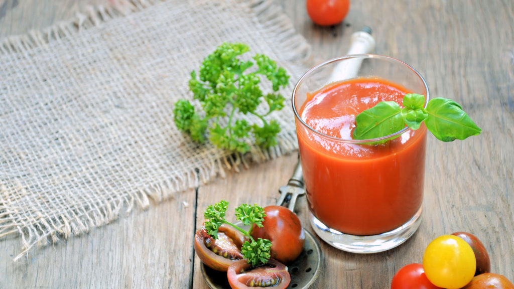 Tomato smoothie in a glass, garnished with fresh basil. Fresh tomatoes and herbs are placed next to it.