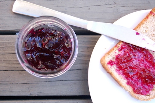 Marmeláda z červených hroznů ve sklenici vedle talíře, na kterém je chléb s marmeládou a nůž.