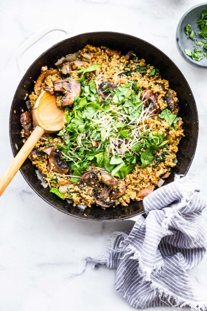 Risotto with millet and mushrooms sprinkled with cheese and leaf spinach in a pan with a wooden spoon.