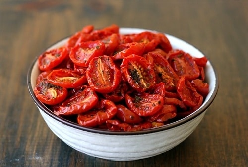 Rozpůlená rajčata, usušená v troubě, ochucená bylinkami a podávaná v bílé misce.