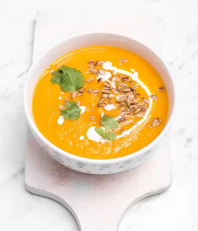 Carrot and pumpkin soup in a bowl on a cutting board decorated with herbs, cream and sunflower seeds.