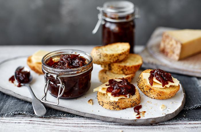 Caramelized onion jam in a glass, which is placed next to sliced ​​bread and a spoon.