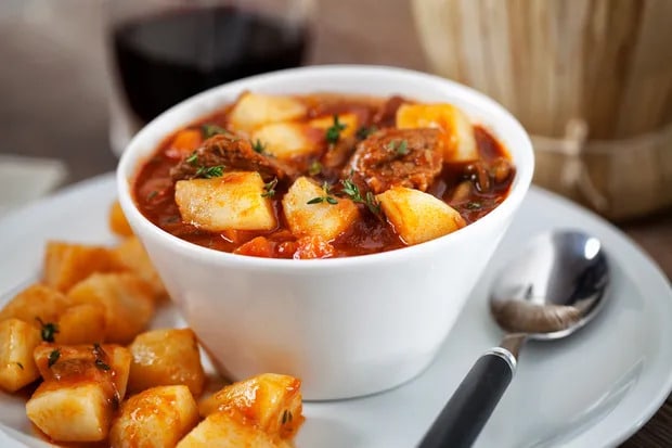 Beef pieces with potatoes and vegetables in a red wine sauce in a bowl with a spoon and potatoes placed next to it.