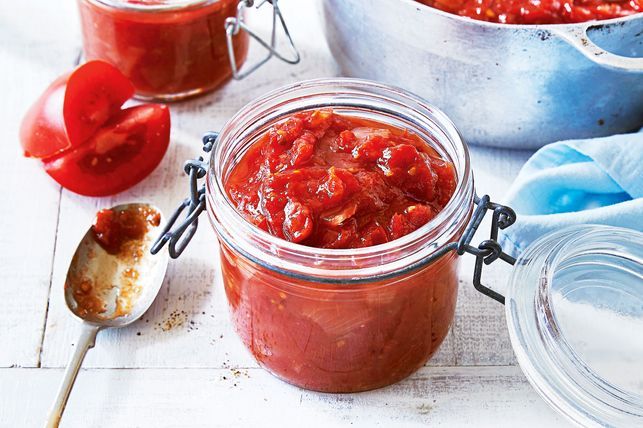 Tomato jam with spices and ginger in a jar with a spoon and tomatoes next to it.