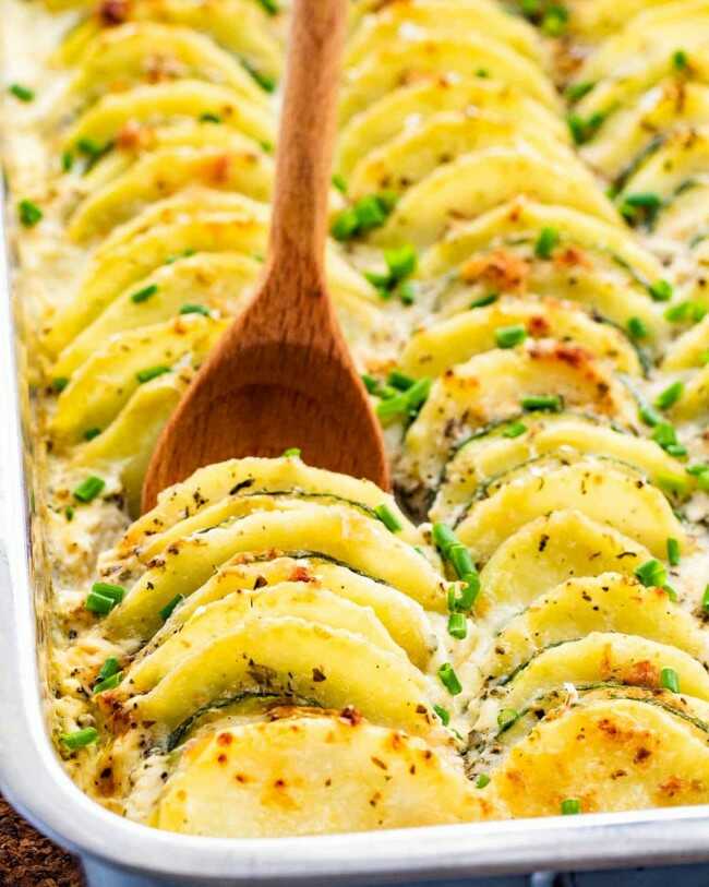 Baked potato slices with zucchini in a baking dish with a wooden spoon.