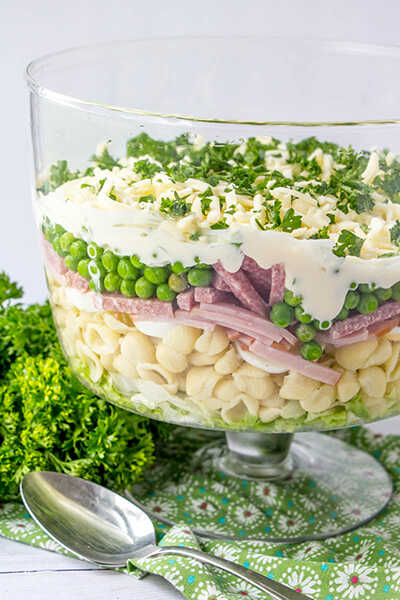 Salat aus Nudeln, Salami, Erbsen, Käse und Dressing bestreut mit frischen Kräutern, serviert in einer Glasschale.