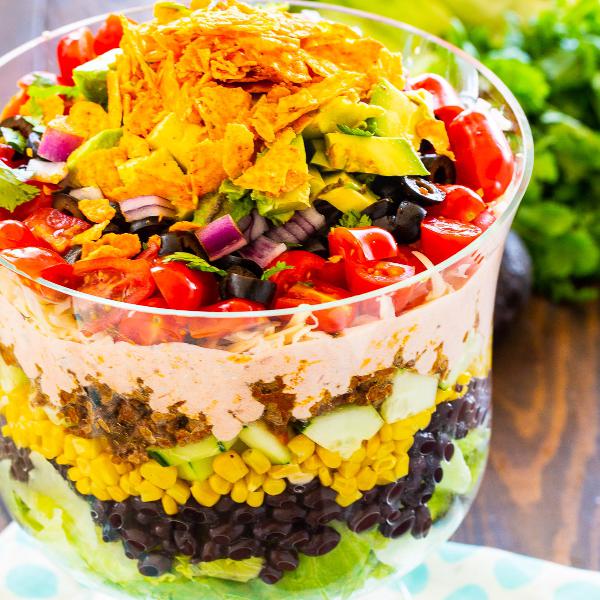 Salad with olives, corn, avocado, tomatoes, red onion and ground meat, sprinkled with nachos and served in a glass bowl.