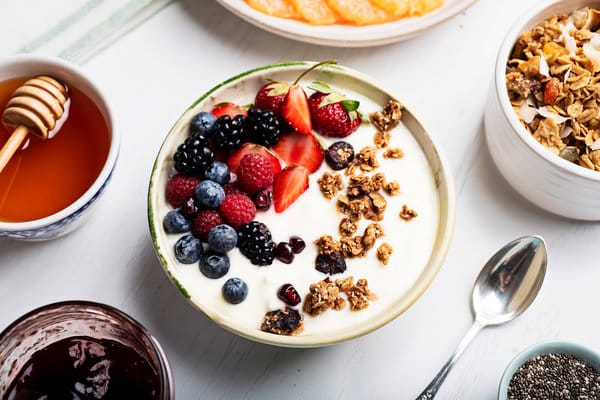 Jogurt vyrobený po domácku servírovaný v misce s müsli a čerstvým ovocem, s vedle položenou lžičkou a mističkami s müsli a sirupy.