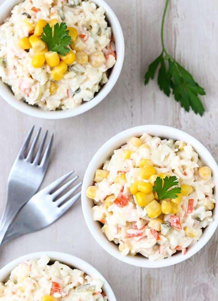 Salat aus Krabbenstäbchen und Reis, serviert in Schalen und garniert mit Mais.