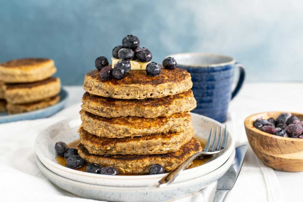 Oatmeal pancakes with seeds served on top of each other on a plate with a fork, garnished with blueberries and butter.