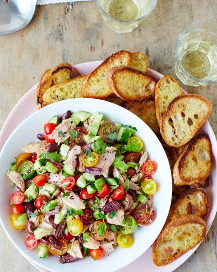 Salát s tuňákem, okurkami, rajčaty, olivami a bylinkami servírovaný na talíří s opečenými plátky bagety.