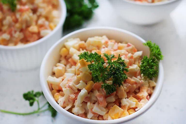 Salat mit Krabbenstäbchen, Mais und Karotten, serviert in Schalen, garniert mit Petersilie.