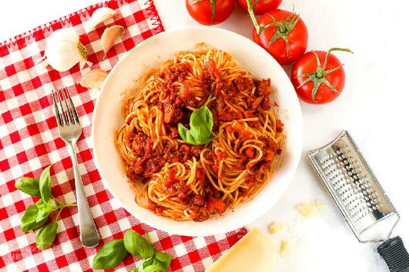 Spaghetti mit Fleisch, Tomaten, Basilikum, Zwiebeln und Karotten auf einem Teller mit einer Reibe, Tomaten, Knoblauch und Basilikum daneben.