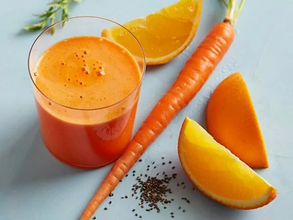 Pomerančovo-mrkvový džus nalitý ve sklenici, která je položena vedle čerstvé mrkve a pomerančů.