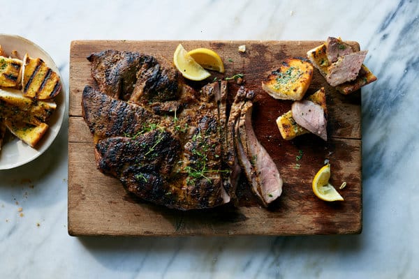 Crispy grilled shoulder seasoned with fennel, coriander and cumin, served on a wooden board with lemon wedges and a side plate of buttery garlic bread.