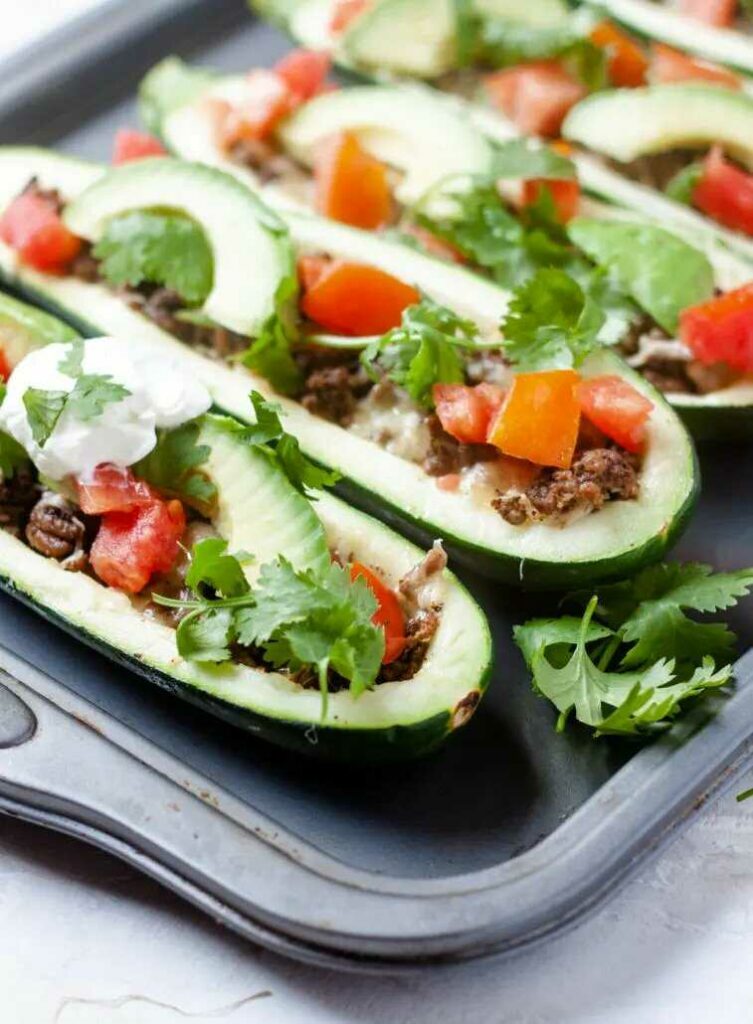 Zucchini boats with minced meat, cheese, fresh tomatoes, avocado and coriander served on a plate.