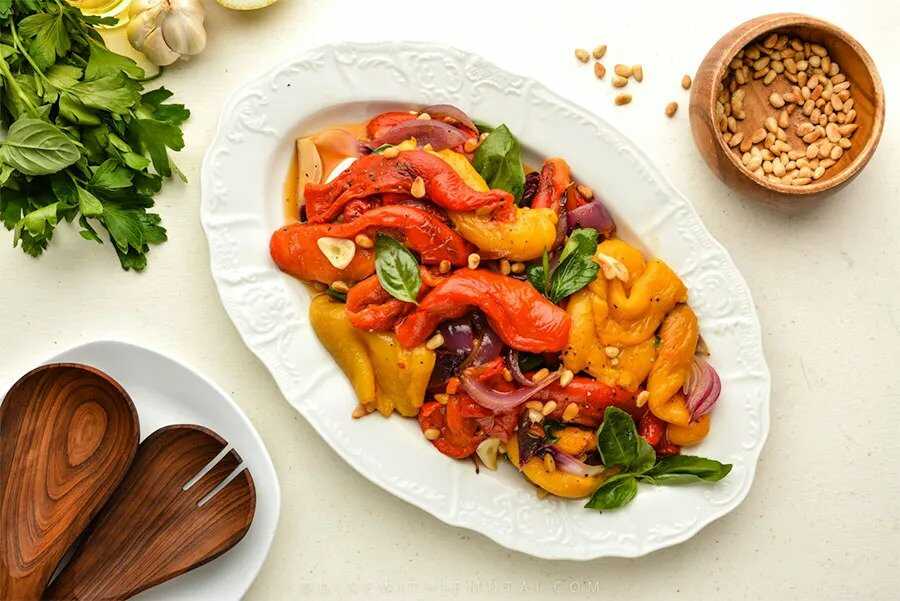 Paprikový salát s restovanou cibulí, praženými piniovými oříšky a čerstvými bylinkami servírovaný na oválném talíři s vedle položenými vařičkami a miskou semínek. 