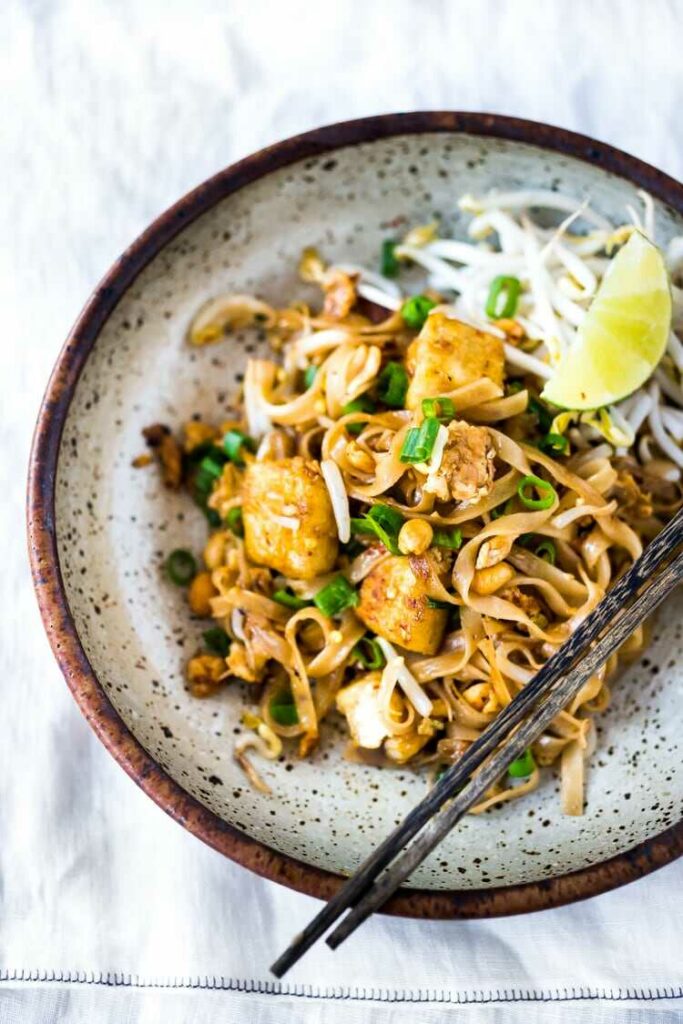 Thajské nudle s tofu, klíčky a lahodnou omáčkou servírované na talíři s čínskými hůlkami a klínkem limety.