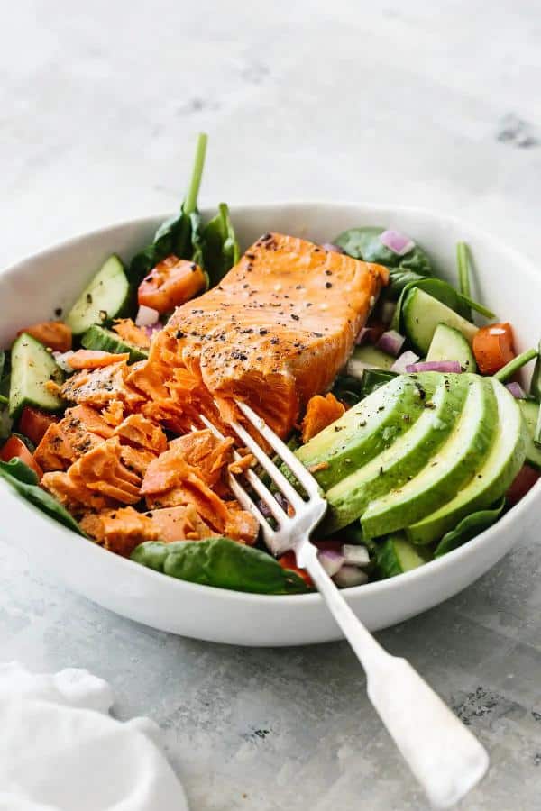 Spinach, avocado, tomatoes and red onions served in a deep plate with a slice of salmon and a fork.