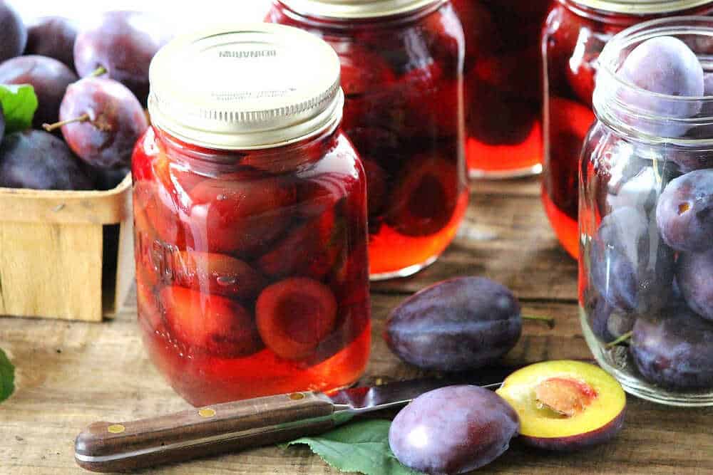 Pickled plums in jars with fresh plums and a knife placed next to them.