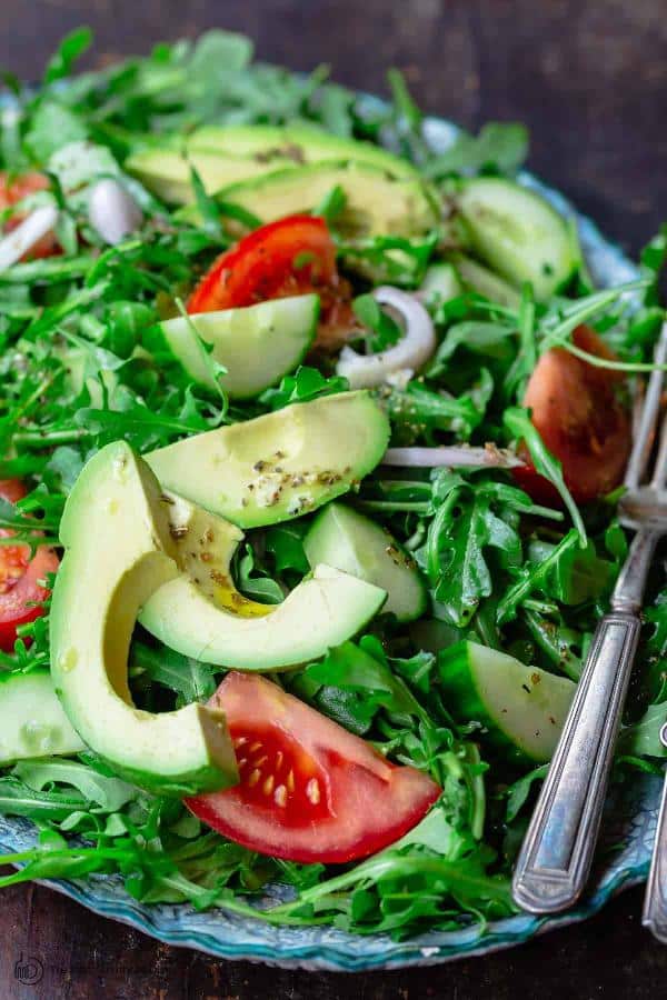 Arugula salad with avocado, tomatoes, cucumbers and shallots, all tossed with a delicious lemon dressing and served in a bowl with a fork.