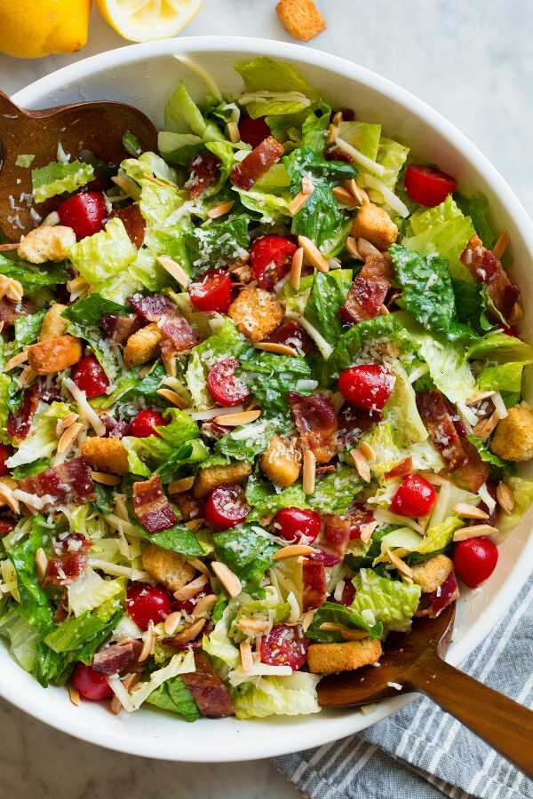 Fresh romaine lettuce, two types of cheese, bacon, cherry tomatoes, roasted almonds and croutons, all tossed with lemon dressing and served in a bowl with wooden spoons.