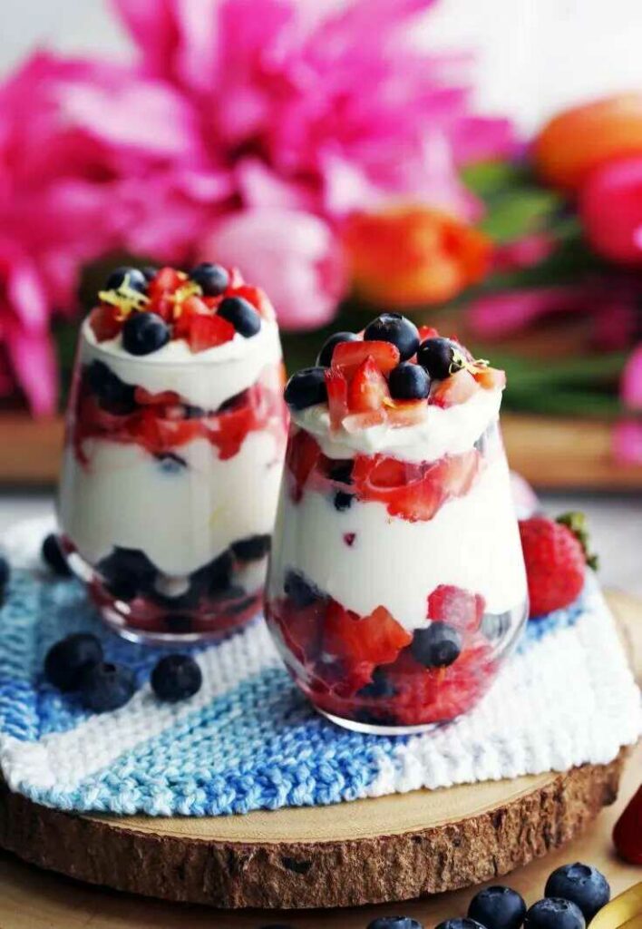 Dvě sklenice s jahodami a borůvkami  v kombinaci s krémem z mascarpone a jogurtu servírované na dřevěném prkýnku s prostíráním.