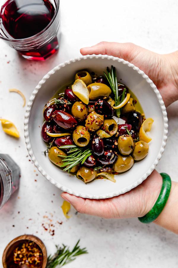Hands holding a bowl full of black and green marinated olives with herbs and a glass of red drink placed next to it.