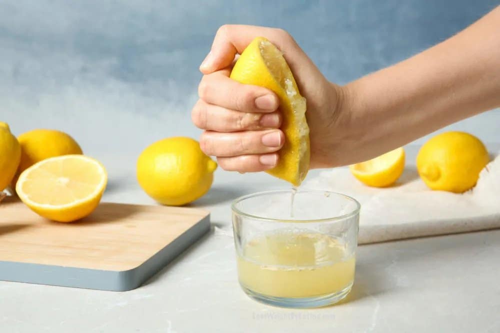 Eine Hand, die eine Zitrone in ein Glas drückt und daneben ein Brett mit frischen Zitronen.