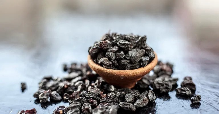 Dried black currants in a small wooden bowl with other dried currants scattered next to it.