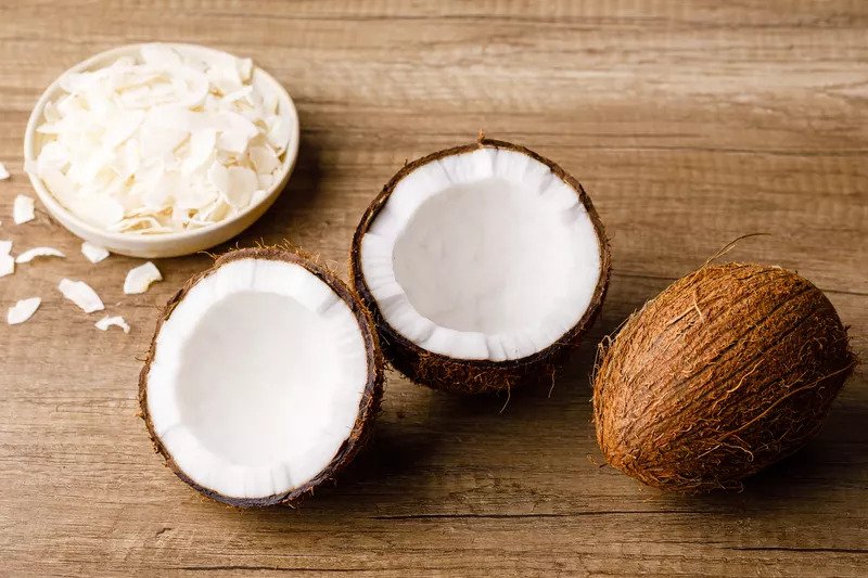 A whole ripe coconut, a halved coconut next to it, and a bowl of coconut flakes next to it.