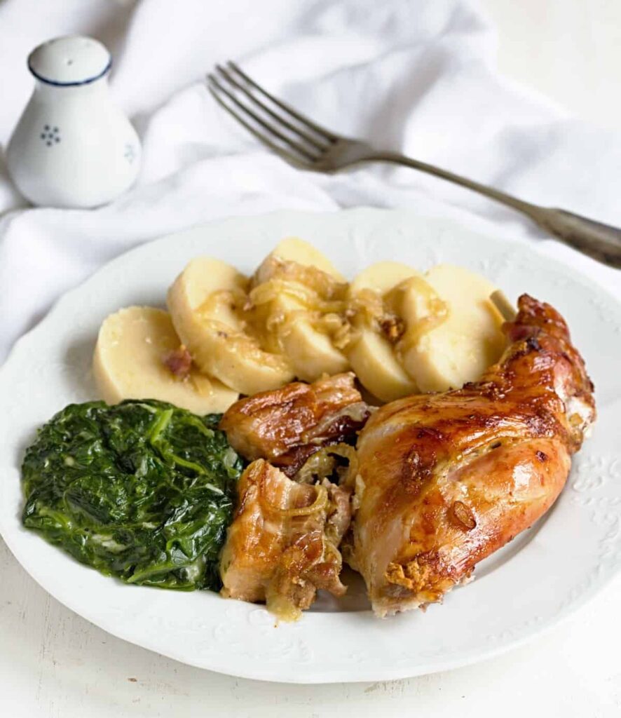 A piece of rabbit on a plate with spinach and potato dumplings with a fork and salt shaker next to it.