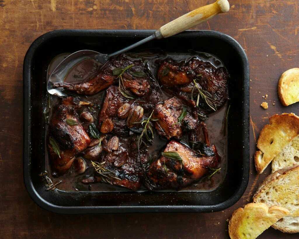 Rabbit in a red wine sauce with fresh herbs served in a roasting pan with a spoon and pieces of toasted bread on the side.