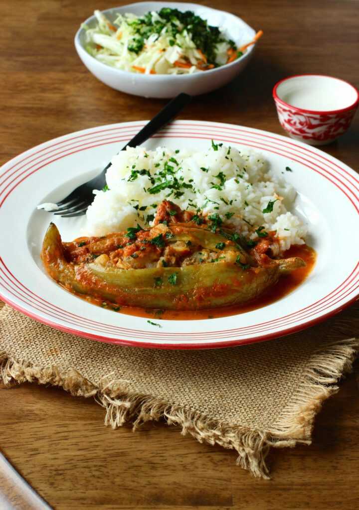 Gefüllte Paprika und Gurken mit Soße und Reis auf einem Teller mit einer Gabel und hinter dem Teller ist ein Teller mit Gemüse.