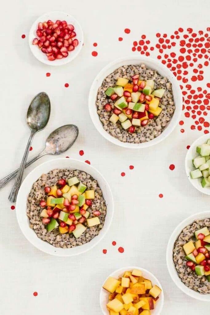Buckwheat porridge served in bowls with fresh fruit. Two spoons, a bowl with a pomegranate and a bowl with a sliced peach are placed next to it.