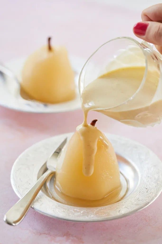 A poached pear on a plate with a fork and a hand pouring sauce over it from a glass.