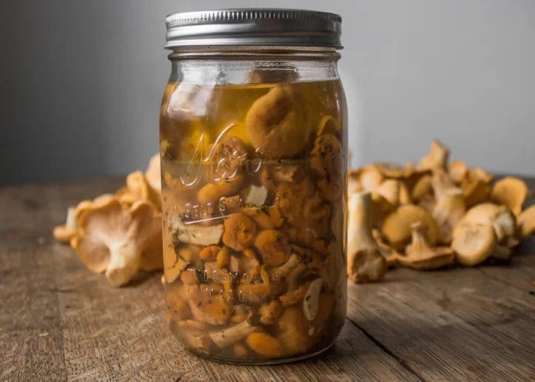 Pickled mushrooms in a mason jar with fresh mushrooms scattered behind it.