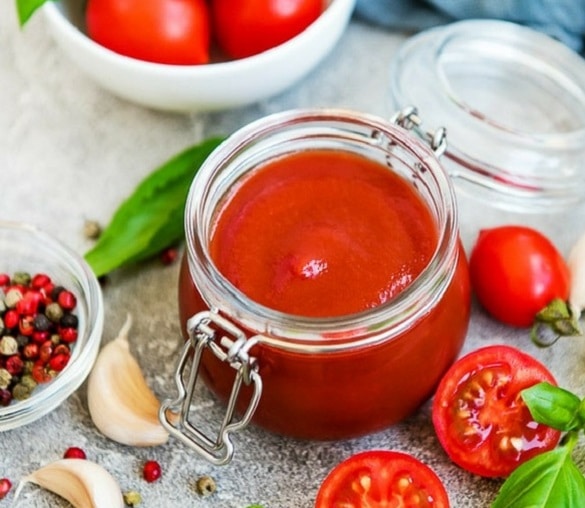 Homemade tomato ketchup in a jar and fresh tomatoes and garlic cloves placed next to it.