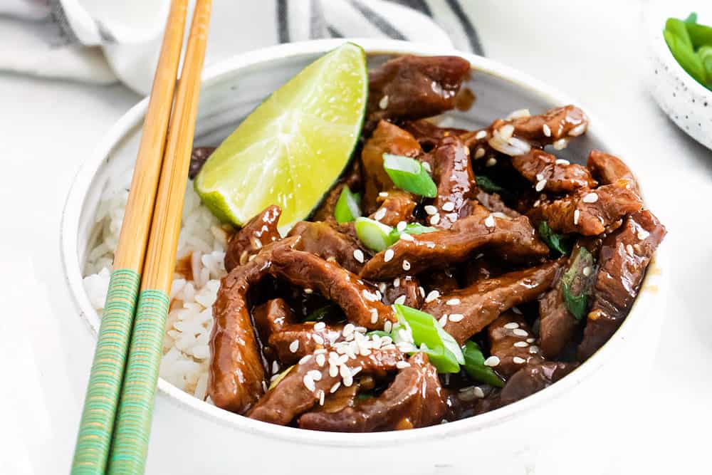 Crispy fried beef in a delicious sweet and spicy soy sauce sauce with fresh ginger, garlic and chilli served in a bowl with rice, lime wedge and chopsticks.