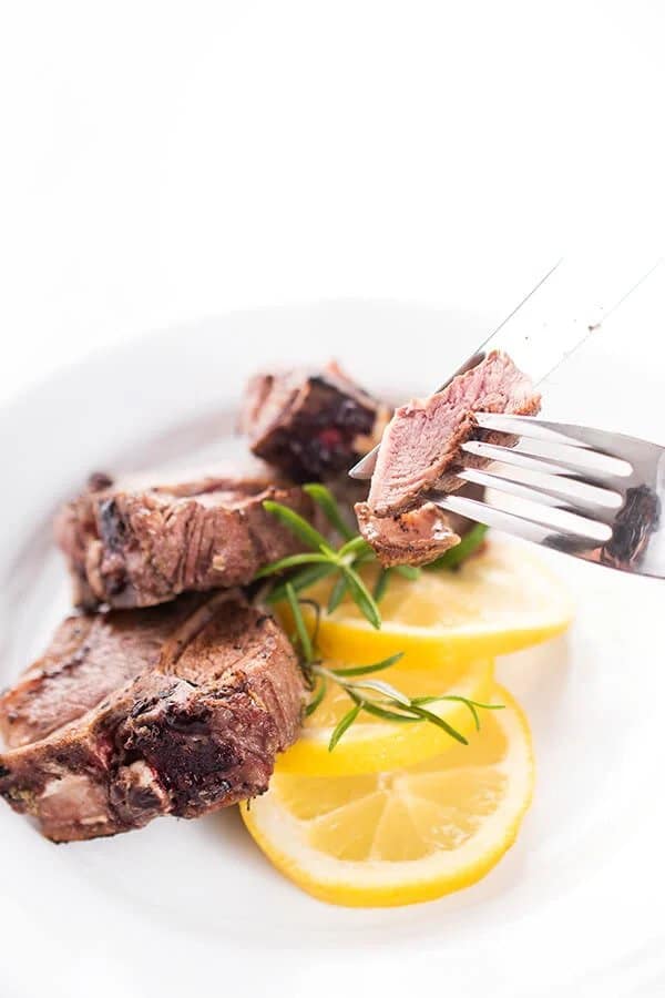 Lamb chops, marinated in a mixture of garlic, pepper and parsley, baked until tender, served on a plate with lemon slices, rosemary and a fork.
