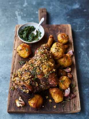 Leg of lamb served on a board with potatoes, garlic and a bowl of spinach with a spoon.