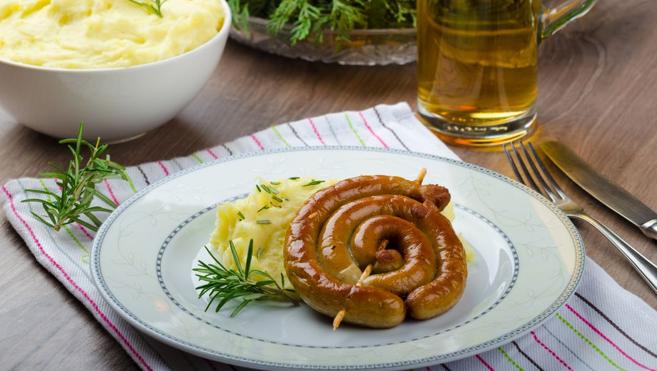 Weinwurst mit Kartoffelpüree serviert auf einem Teller mit einem Glas Bier, Besteck und einer Schüssel Kartoffelpüree daneben.