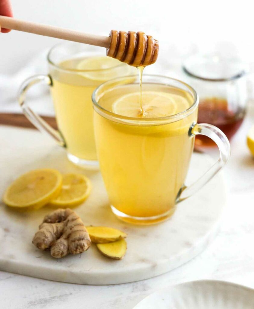 Čaj ze zázvoru ve skleněných hrníčcích s plátky citronu a vedle je položen kus čerstvého zázvoru a další plátky citronu.