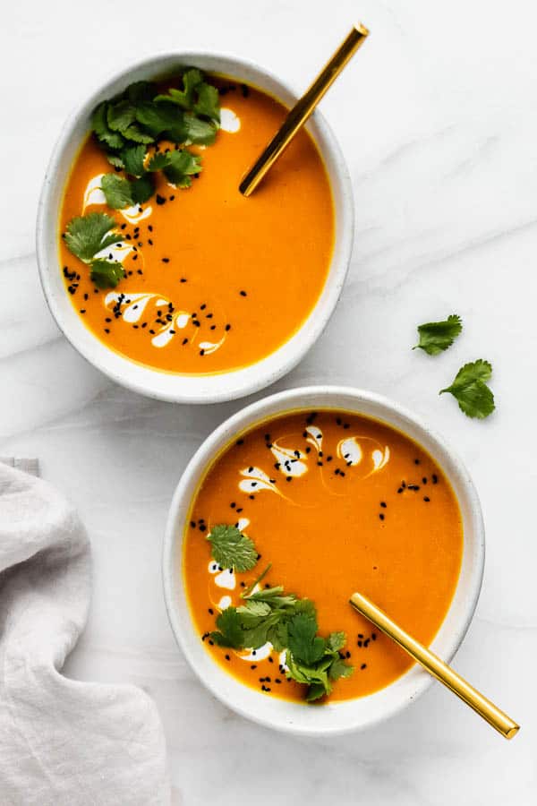 Carrot, ginger and curry soup served in two bowls with straws, garnished with fresh herbs, cream and seeds.
