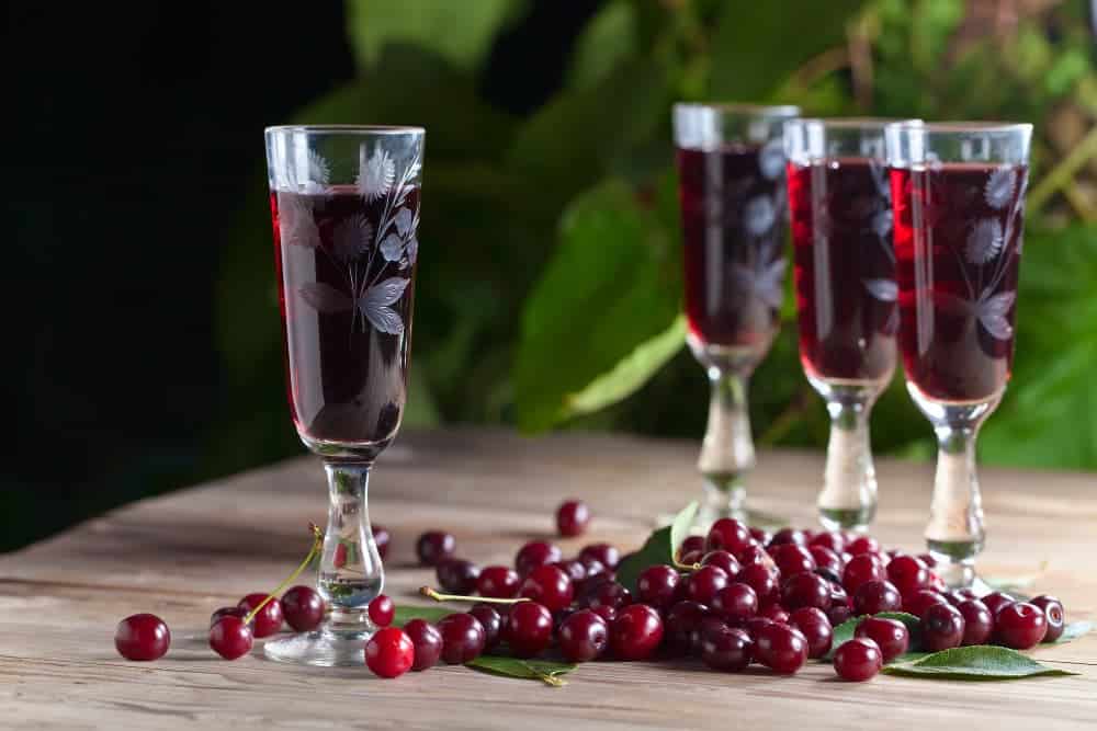 Domácí likér z višní rozlitý ve sklenicích s vedle rozsypanými čerstvými višněmi.