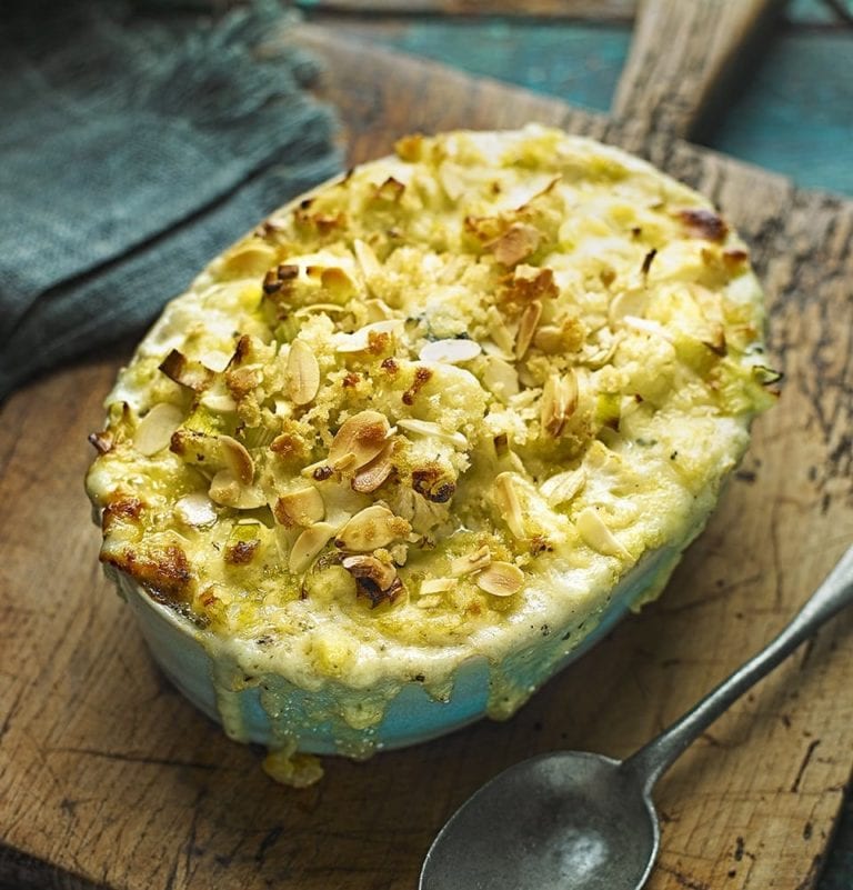 Cauliflower gratin with cheese and leek, sprinkled with almonds and served in a baking dish with a spoon on the side.